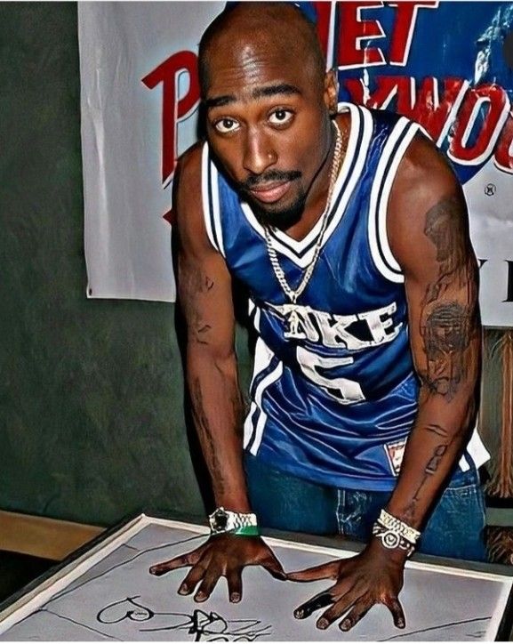 Tupac vestindo uma camisa de basquete azul e olhando fixamente para a câmera