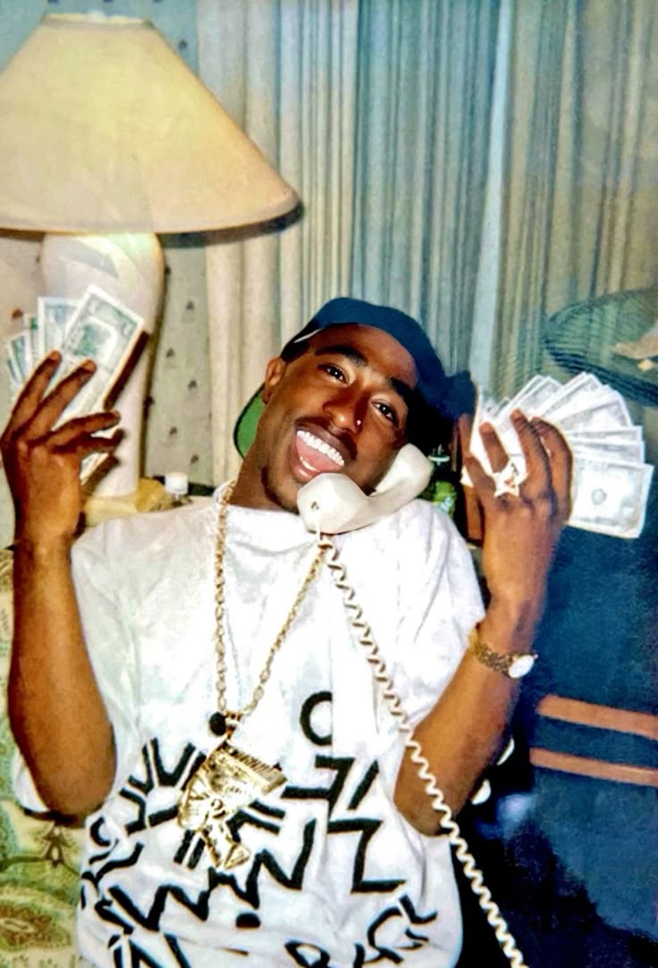 Tupac sorrindo, falando ao telefone e segurando maços de dinheiro